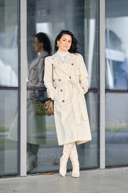 A woman showcases her autumn street style, wearing a cream coat and boots while leaning against a glass building. The scene reflects a stylish urban background, perfect for a fall day