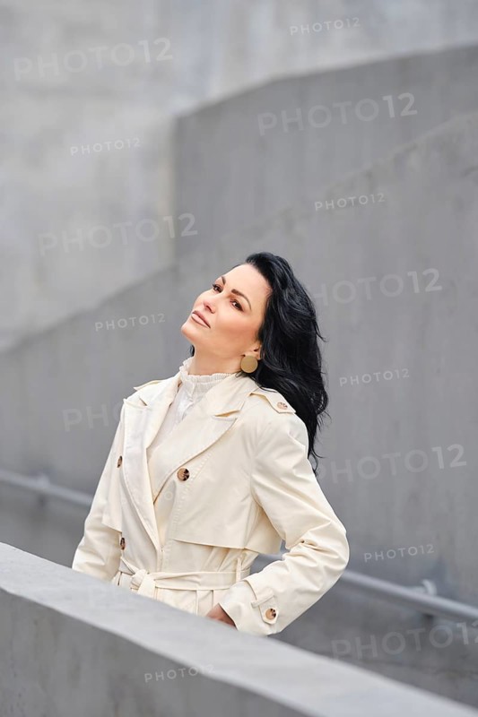 A woman stands confidently in a fashionable trench coat, gazing upward. The backdrop features sleek architectural lines. It's a crisp autumn day, capturing the essence of seasonal street style