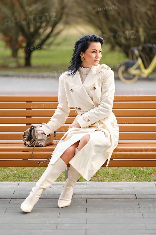 A woman dressed in a stylish cream trench coat and knee-high boots sits gracefully on a wooden bench in a peaceful park