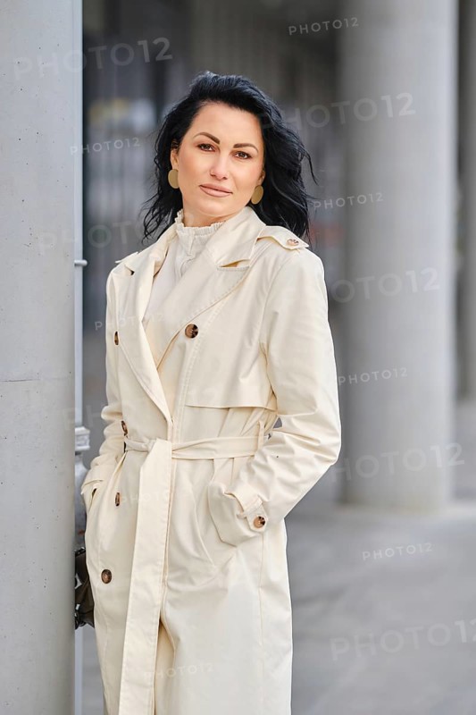 An urban portrait of a woman in a cream trench coat . The scene showcases autumn weather with a city backdrop