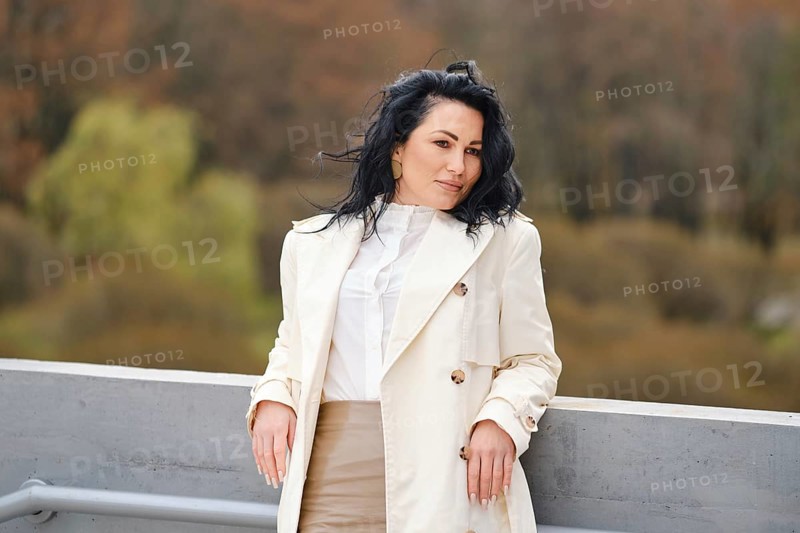 Smiling woman stands against a backdrop of autumn foliage, wearing a light trench coat and stylish outfit. The warm colors of fall enhance her fashionable street style in this serene location