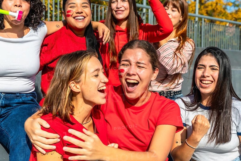 Group of excited diverse young women fans cheering loudly and celebrating a sports victory or game while sitting on stadium bleachers with red face paint