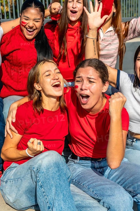 Group of enthusiastic women wearing red shirts and face paint, celebrating with open mouths and raised fists while watching a sporting event from the stadium stands