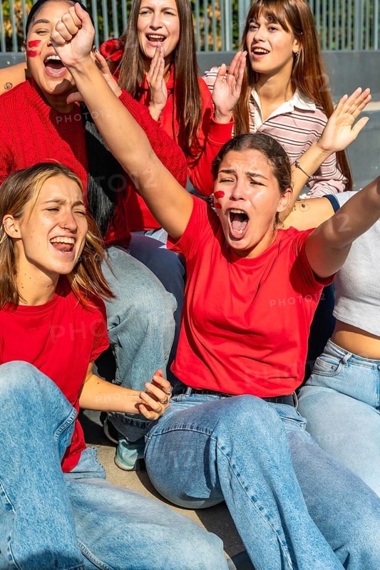 Young women sitting together, wearing red shirts and face paint, passionately cheering and celebrating their team's performance with arms raised and open mouths expressing pure excitement
