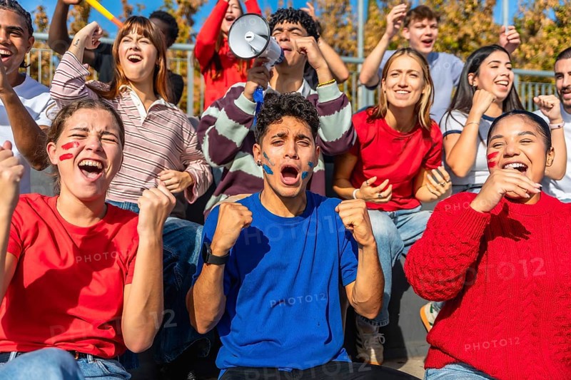 Excited sports fans yelling and cheering with painted faces, pumping fists, and using a megaphone while watching a game from stadium bleachers, sharing a moment of triumph and community