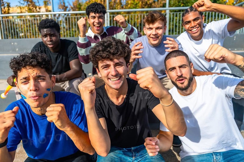 Enthusiastic young diverse male friends sitting on grandstand steps watching a sports event, celebrating intensely with raised fists and excited expressions