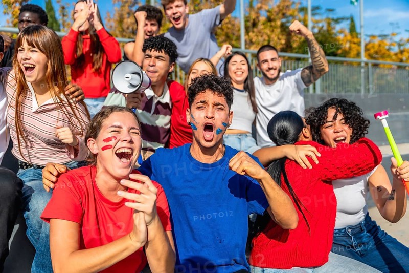 Group of diverse young sports fans celebrating and cheering loudly, showing strong emotion with painted faces, holding a megaphone and a vuvuzela while watching a game at an outdoor stadium