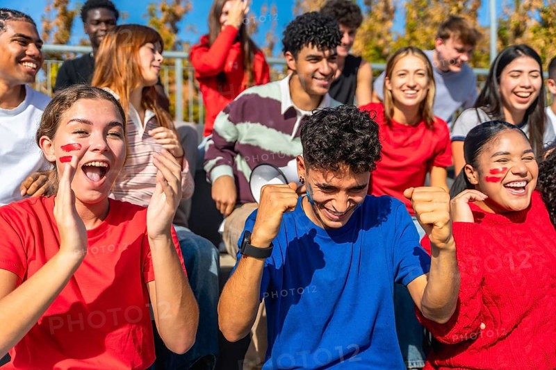 Energetic, diverse group of young adult spectators passionately cheering and celebrating their team's victory at a sports event in an outdoor stadium, showing excitement and unity