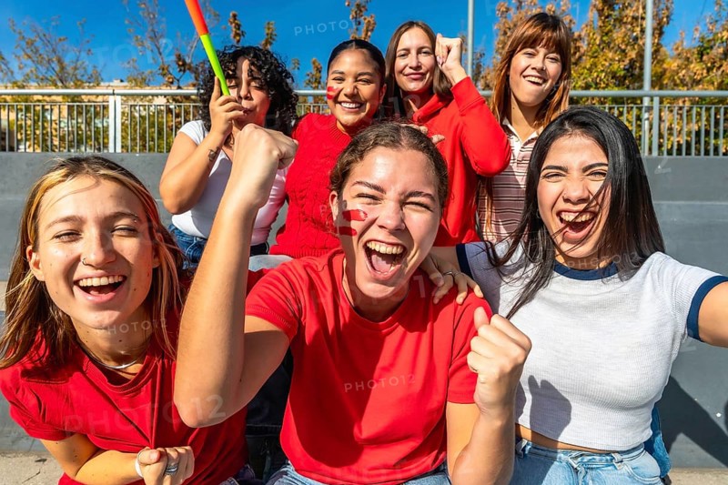 Diverse group of young women cheering loudly in a stadium, faces painted and fists raised, celebrating their team with joy, energy and friendship during a daytime game