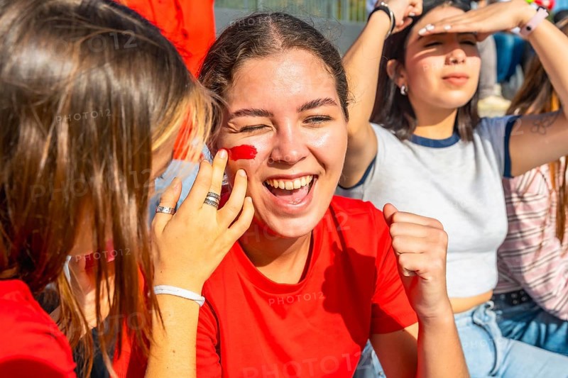 Young woman with red face paint cheering joyfully and raising her fist, celebrating team victory with friends at a stadium while expressing excitement and happiness