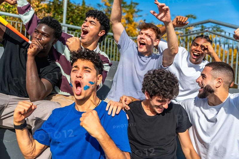 Diverse group of young men with painted faces and horns celebrating loudly, showing excitement and support during a thrilling sports event in a stadium