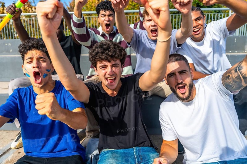 Excited multiethnic male friends cheering and raising fists in outdoor stadium, standing together in bleachers, smiling, shouting and celebrating their teams winning moment