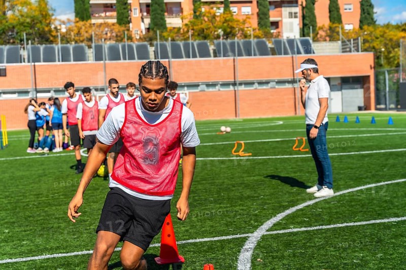 Young male soccer player in a red bib practices drills on green artificial turf with teammates while coach supervises, focusing on technique, movement and teamwork during training