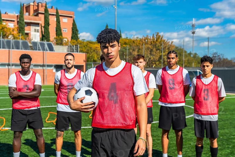 Multi ethnic group of young men in red bibs standing on a green football pitch, preparing for training or a match while showing teamwork and determination