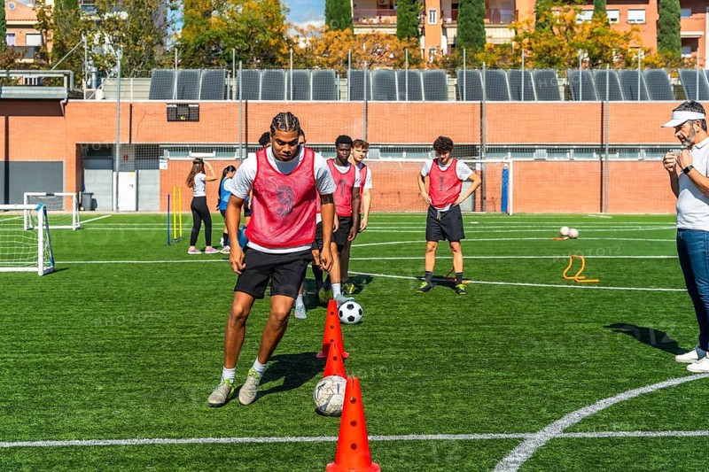 Young men in red vests are performing agility drills with cones and soccer balls on a green artificial turf field, receiving instruction from their coach