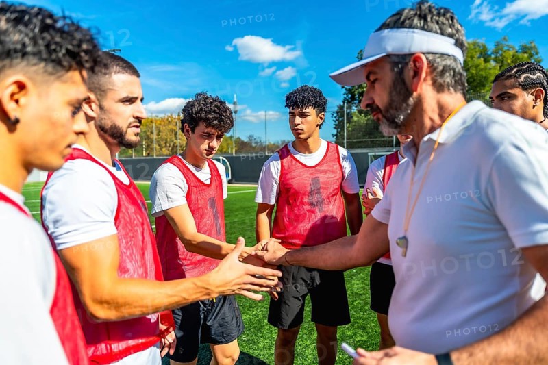 Soccer coach motivating and encouraging young male players with a handshake on a training field under a clear blue sky, fostering sportsmanship and teamwork