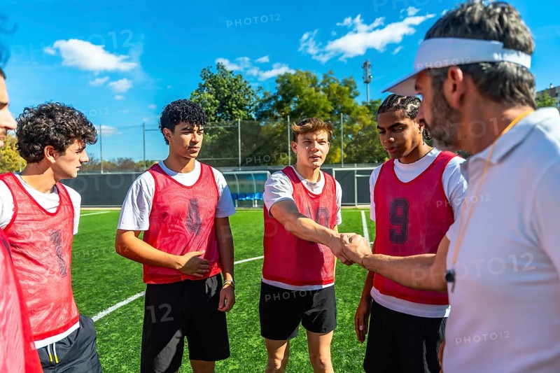 Coach wearing a visor and whistle shaking hands with a young male soccer player on a green football field, with teammates watching during a training session