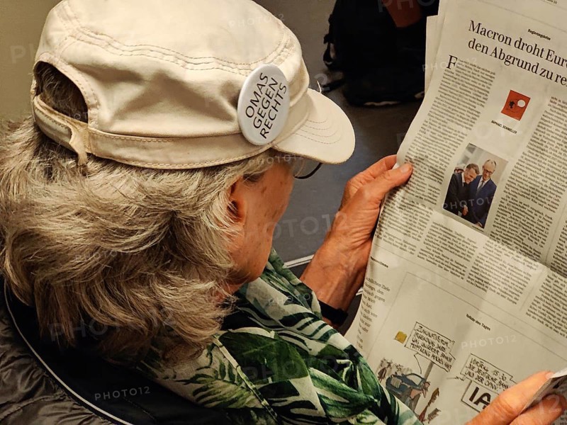 Elderly woman reading in the newspaper with a sticker on her cap from the cross-party citizens' initiative Omas gegen Rechts, Essen, Germany