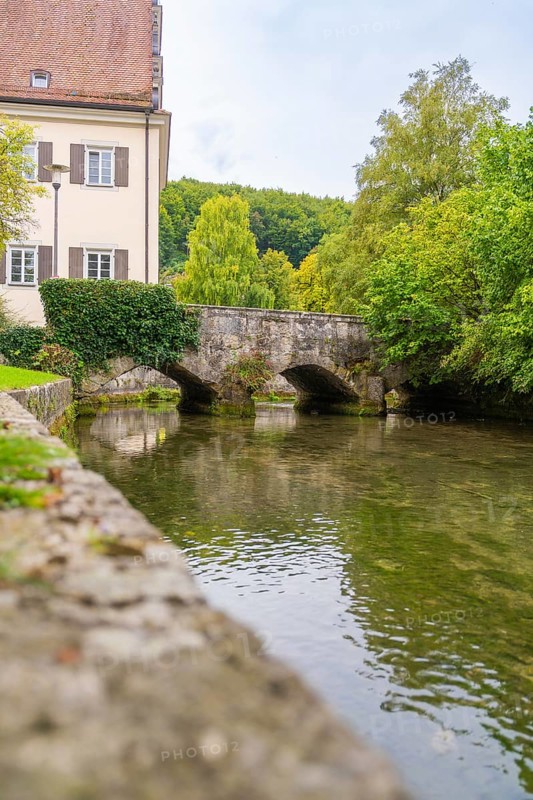 An old stone bridge over a quiet river flanked by buildings and trees, ZFP Zwiefalten, Reutlingen district, Germany