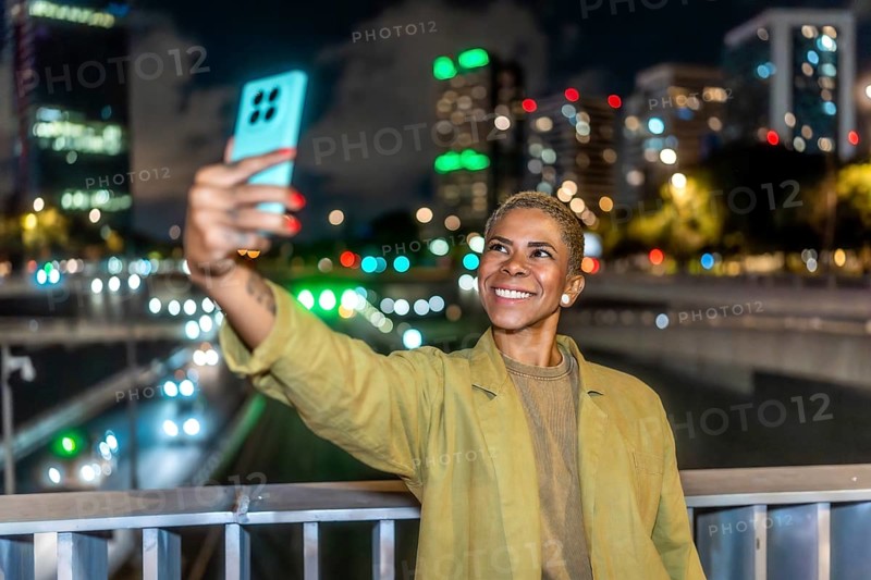 Woman with short blonde hair and happy expression capturing a selfie with her smartphone against a vibrant urban cityscape at night, showcasing technology and connection after dark