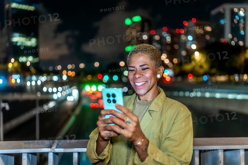 Woman smiling while interacting with her mobile phone, standing on a bridge with blurred city lights and traffic creating a bokeh effect in the urban background at night
