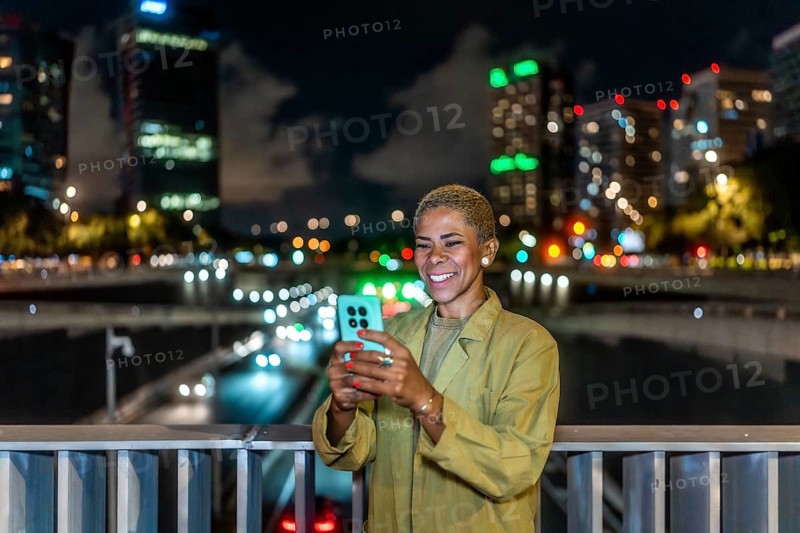 Woman standing on a city bridge at night, smiling while using a smartphone with blurred traffic and illuminated buildings creating a warm bokeh background