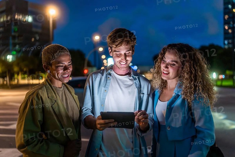 Three diverse friends, two women and one man, standing on a city street at night, smiling and enjoying social connection while looking at content on a shining tablet