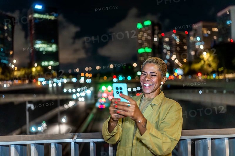 Happy woman smiling, holding a smartphone and enjoying a vibrant urban night view from a bridge, with illuminated city buildings and bokeh lights in the background