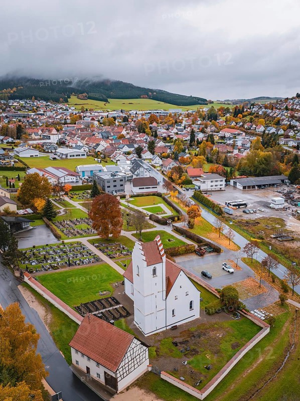 View of a town with church and cemetery surrounded by autumn trees and residential buildings on a cloudy day, Wehingen, Tuttlingen district, Germany