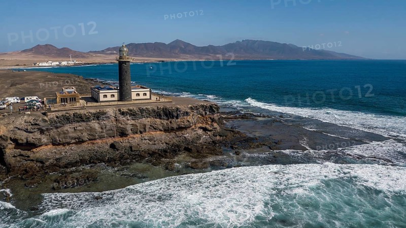 Aerial view of Faro Punta de Jandia lighthouse at the southern tip of Jandia peninsula, in the foreground white foam crowns on moving sea swell large high waves on reef edge beaming sea strong surf, Fuerteventura, Canary Islands, Canary Islands, Spain