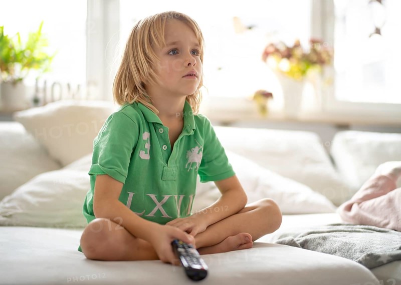 Child, boy, 7 years old, blonde, sitting on sofa, with remote control in hand, watching TV, watching television, watching television, excited, fascinated, Stuttgart, Baden-Württemberg, Germany