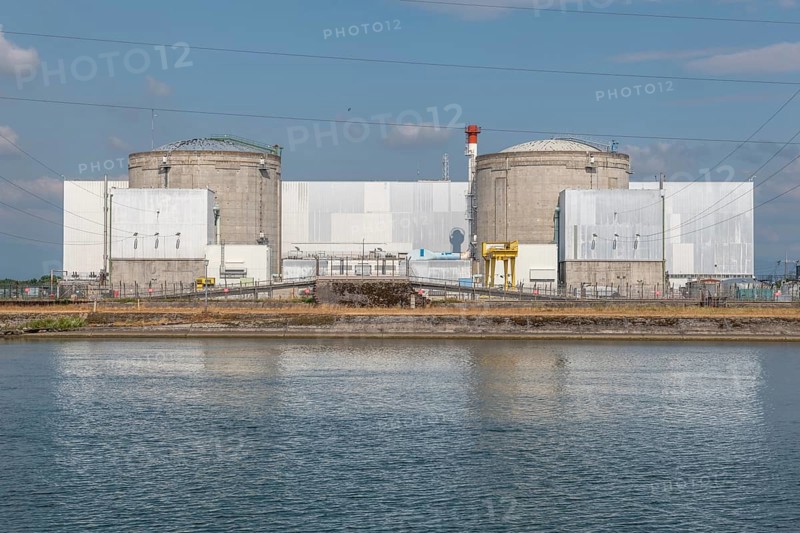 A large industrial nuclear power plant is located on a quiet water front. Two tall cylindrical structures dominate the skyline, with overhanging power lines and distant clouds that ennoble the blue sky. Fessenheim, Haut Rhin, Alsace, France