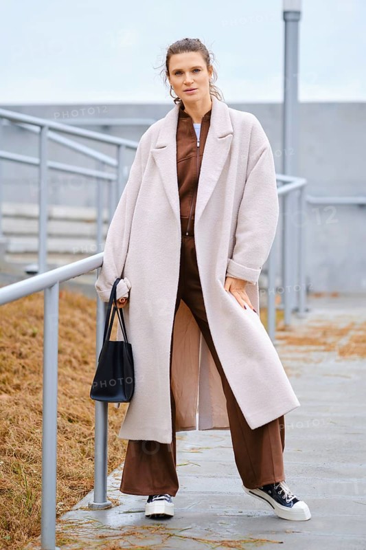 A woman stands confidently on outdoor steps, wearing a long light coat over a brown outfit. She carries a black bag and wears sneakers, with a gray sky visible above her