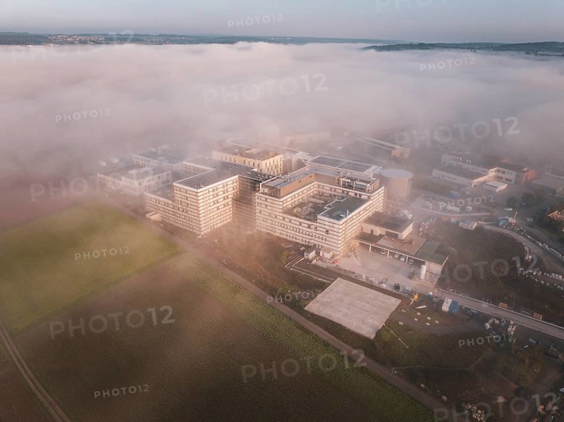 Large industrial buildings surrounded by fields under a thick layer of fog, new hospital health campus building, Calw, Germany