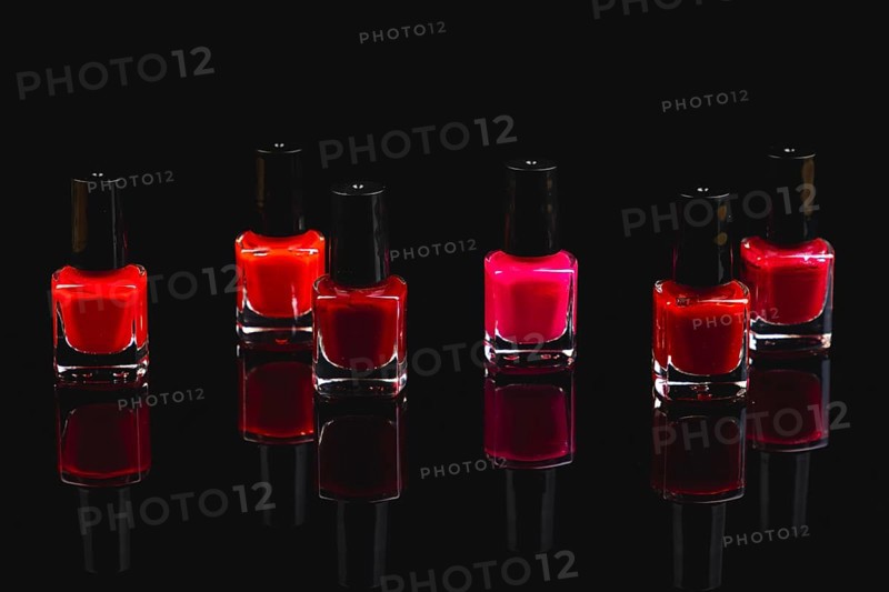 Six bottles of nail polish in shades of red and pink on a black surface with reflection