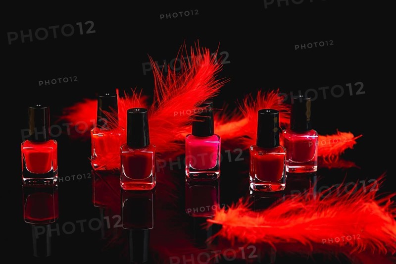 Nail polish bottles in shades of red and pink with red feathers on a black surface with reflection