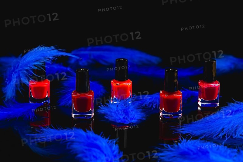Red nail polish bottles surrounded by blue feathers on a black surface with reflection