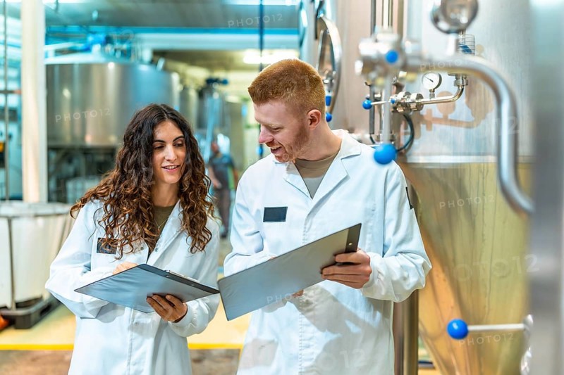 Two brewery workers in lab coats inspecting machinery and documenting processes, ensuring product quality and efficient operation in a modern industrial setting
