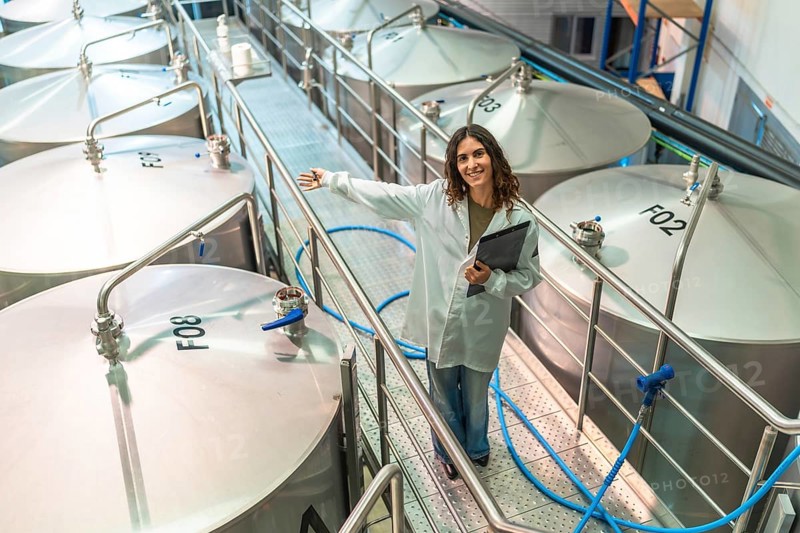 Woman wearing a white lab coat and holding a clipboard, standing on a metal gangway, showing a row of large stainless steel fermentation tanks in a modern brewery