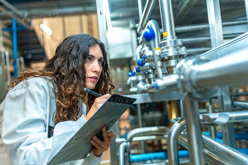 Woman in a white coat diligently checking and documenting the status of stainless steel pipes and valves inside a modern food and beverage production facility