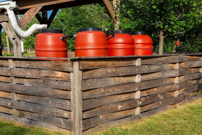 Reddish brown plastic water butts collecting and storing rain water from white plastic downspout in community garden enclosure in summer, Quebec, Canada