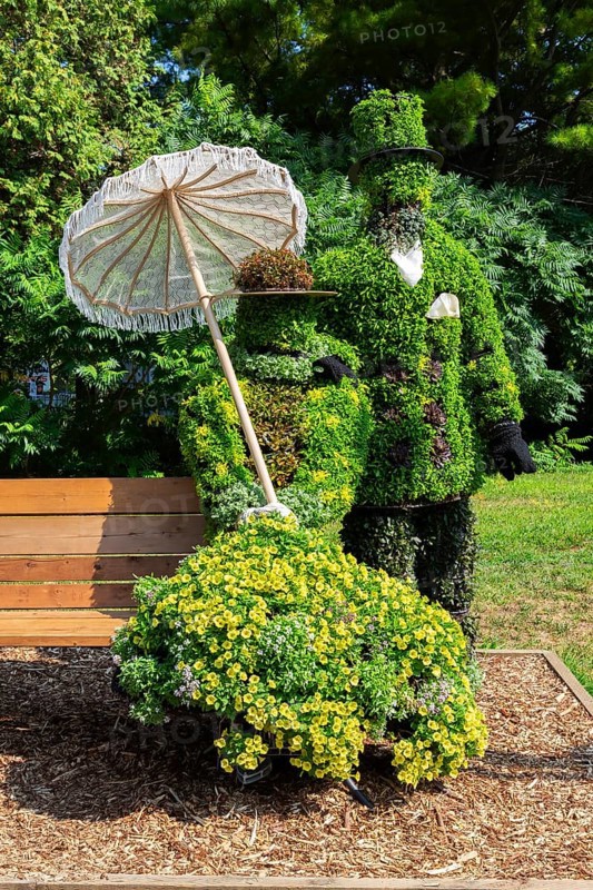 Live plant sculpture of Victorian man standing and woman with sun shade umbrella sitting on wooden park bench in summer, Ile des Moulins, Old Terrebonne, Quebec, Canada