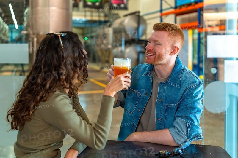 Young couple on a brewery date tasting craft beer, smiling and relaxed in a modern industrial taproom, enjoying pints, conversation and a cheerful weekend outing