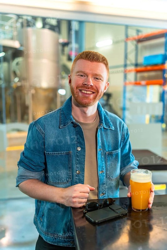 Smiling man enjoying a glass of craft beer in a modern industrial brewery. He stands wearing a denim jacket, looking at the camera with a happy expression