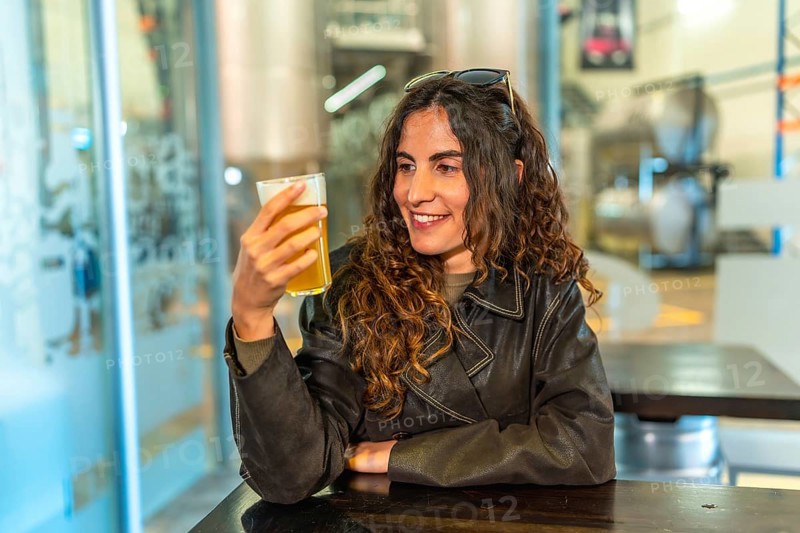 Woman in a leather jacket smiling and savoring a glass of craft beer at a modern brewery bar, enjoying a relaxed tasting experience with friends in a casual weekend hangout