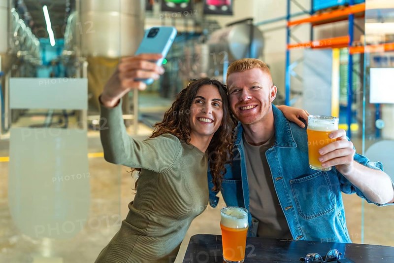 Young couple smiling for a selfie with craft beers in hand at a modern brewery tasting room, enjoying a casual weekend hangout, tasting and sharing the experience