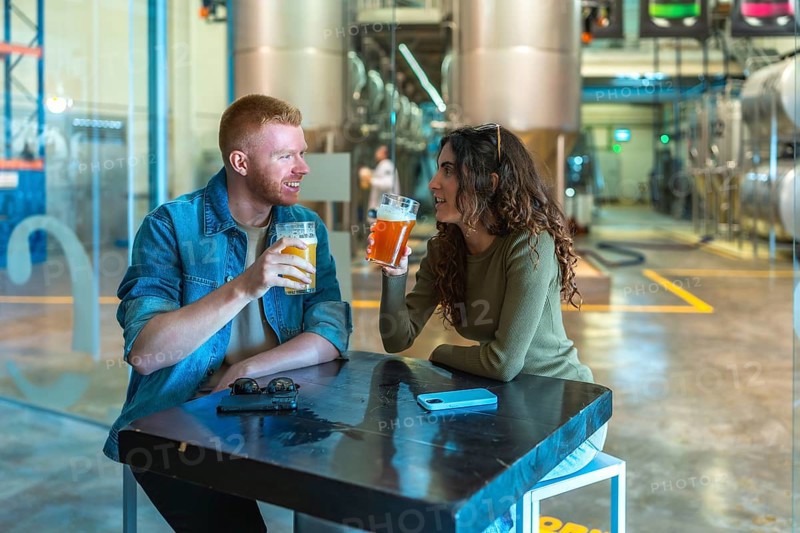 Couple sitting at a table in a modern craft beer factory, drinking and communicating while enjoying a brewery tour experience with large fermentation tanks in the background