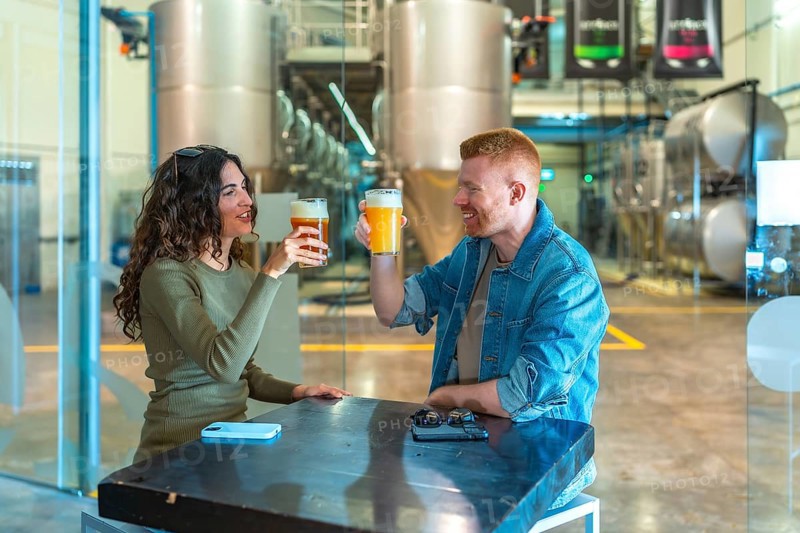 Young couple enjoying craft beer at a table, making a toast and smiling inside a modern beer factory with large stainless steel tanks in the background