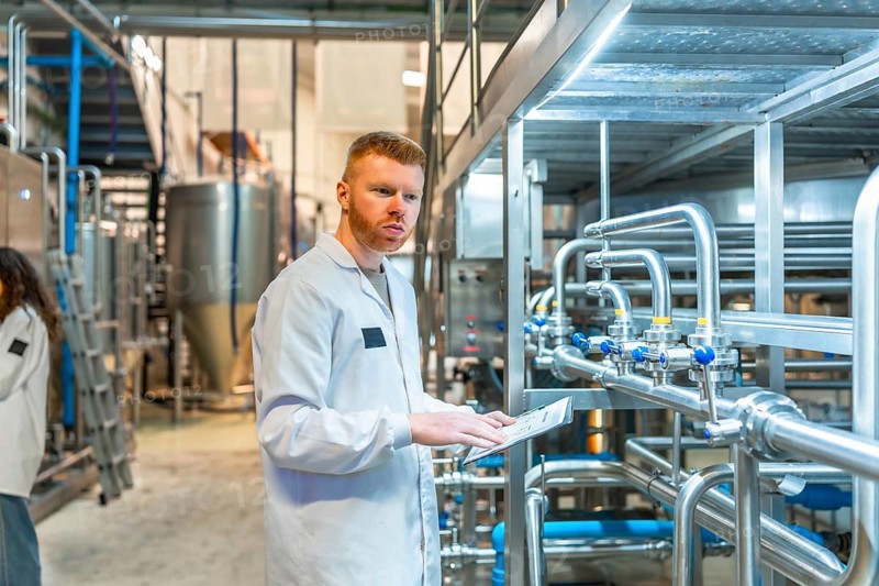 Young male specialist wearing a white lab coat and observing pipes and machinery while holding a clipboard, ensuring quality control and efficient operation in a brewery
