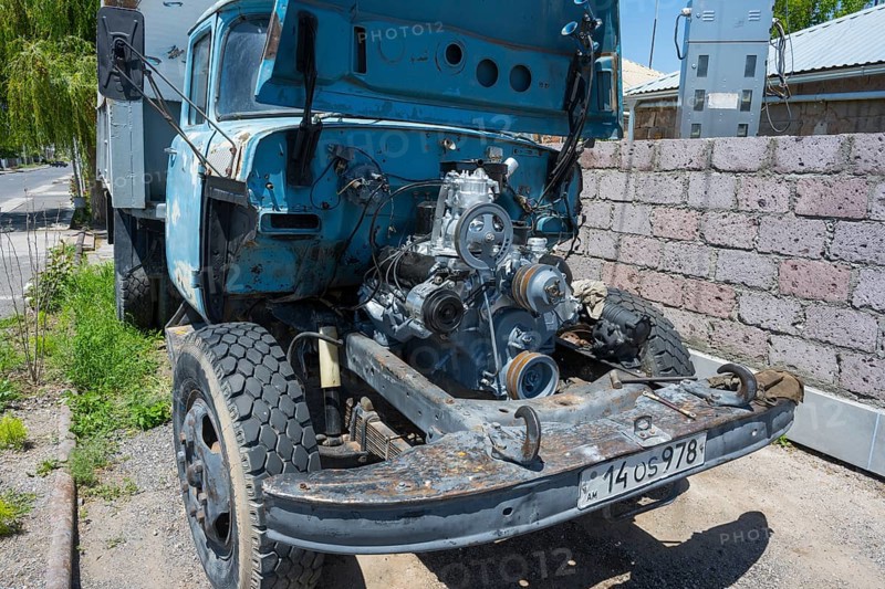 An old lorry with an open bonnet and no front body parts stands close to a brick wall in the sun, engine repair, Armenia